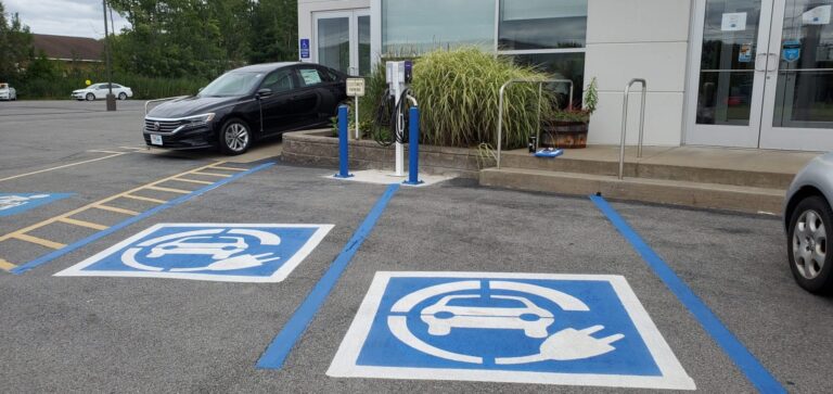 Work done by the Green Team. A designated electric vehicle charging area in front of a commercial building, featuring clear EV parking symbols, a dual-port charging station, and a parked black sedan.