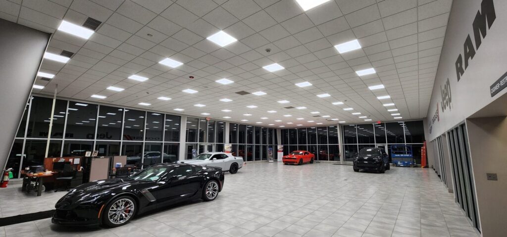 Work done by the Green Team. A spacious car show room with multiple cars illuminated by lights in grid ceiling.