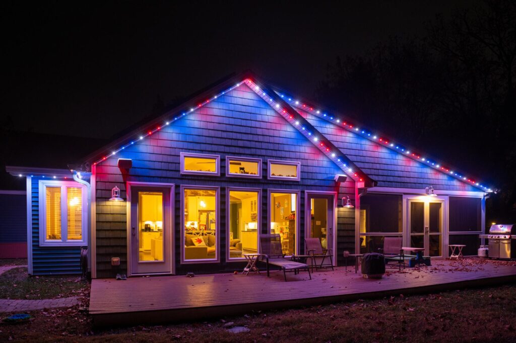 A house illuminated with Oelo permanent holiday lighting in red, white, and blue colors, showcasing a festive design. The lights are installed along the roofline, blending seamlessly with the house exterior at night