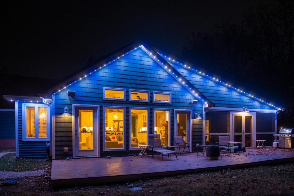 A house illuminated with Oelo permanent holiday lighting in white, and blue colors, showcasing a festive design. The lights are installed along the roofline, blending seamlessly with the house exterior at night