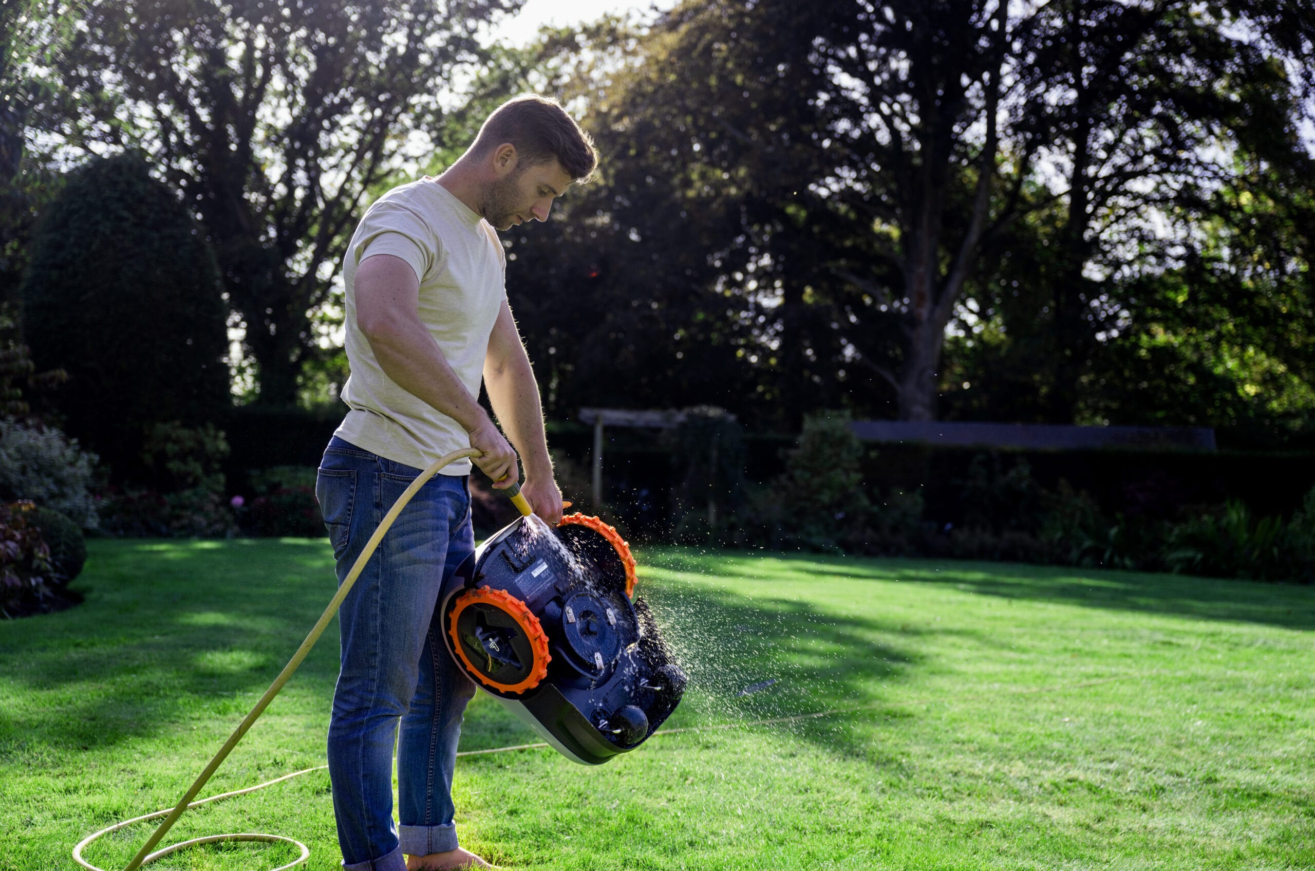 Man cleaning a Segway Navimow robotic lawn mower with a garden hose on a sunny lawn.