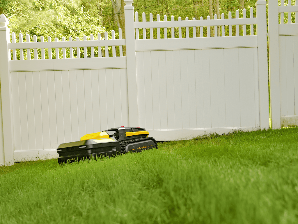 Yarbo Pro Mower trimming grass near a white vinyl fence on a sunny day.