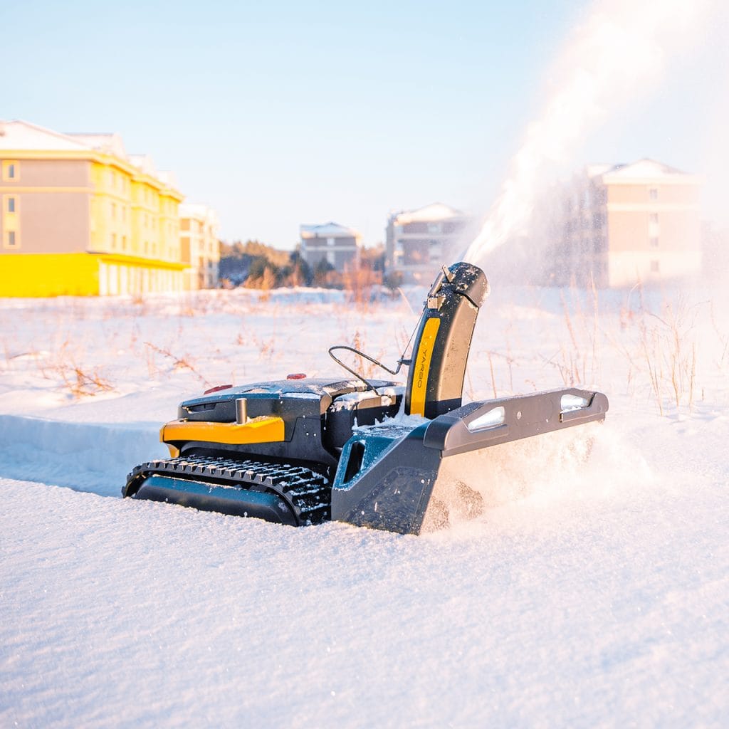 Yarbo robotic snow blower clearing deep snow in an open outdoor area with buildings in the background.